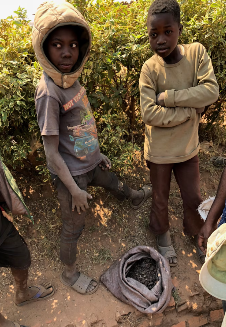 طفلان واقفان قرب كيسهما الذي يحتوي نثرا من الكوبالت. ينخرط الاف الاطفال في تعدين الكوبالت في الكونغو مع عدم قدرة أسرهم على دفع أقساط تعليمهم بسبب ضألة مداخيلها
