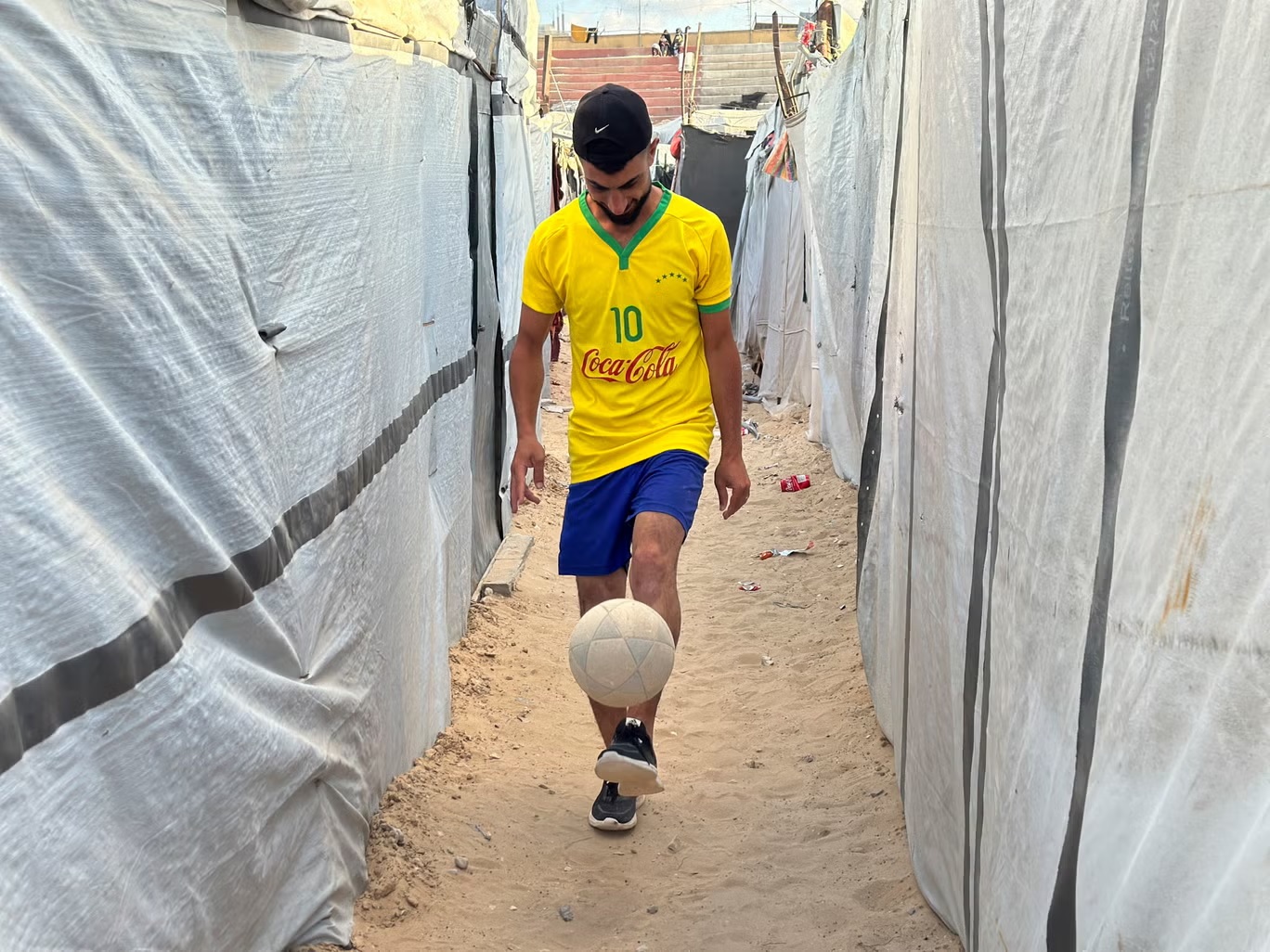 لاعب كرة قدم فلسطيني يراوغ بالكرة بين خيام تؤوي فلسطينيين نازحين في "نادي خدمات خان يونس" - الذي تعرض لأضرار جراء الحرب - في "خان يونس"، قطاع غزة، 22 يونيو 2026 (أ ف ب/ غيتي)